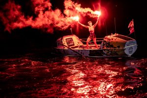 A sailor holds a lit flare at night after finishing an Atlantic crossing.