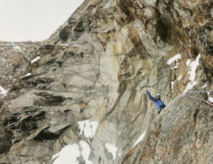 A climber cheers from a vertical granite face, with some patches of snow.