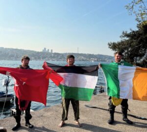 Three paddlers holding flags stand on shore in celebration