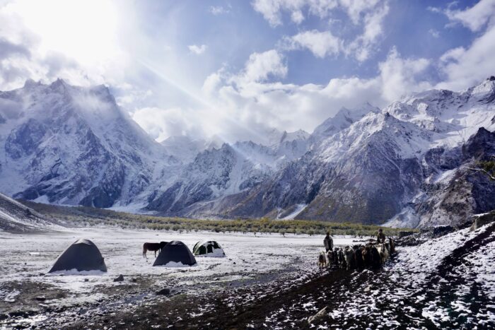 A tent on a snow-powdered plain, with a sheppher and goats on one side, and jagged snowy peaks on the other. 