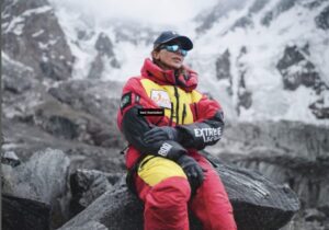 Naila Kiani sits on a rick sourrounded by mountains covreed in clouds, and dressed in a dawnsuit.