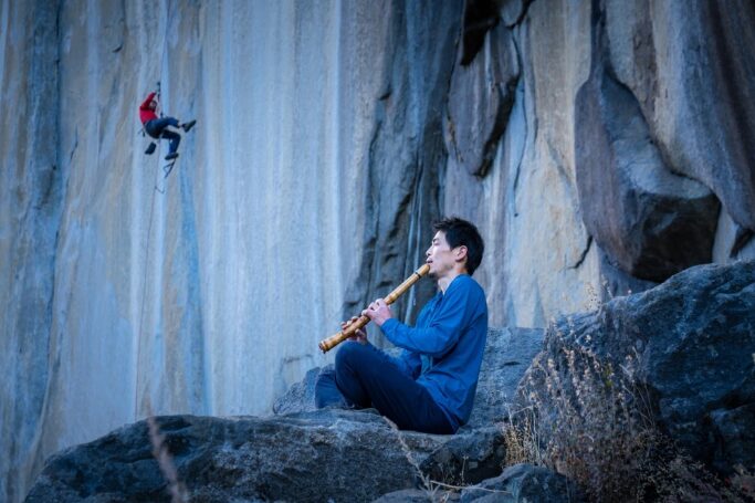 Keita Kurakami playing the flute sitting on a pad, a climber behind on Yosemite's walls.