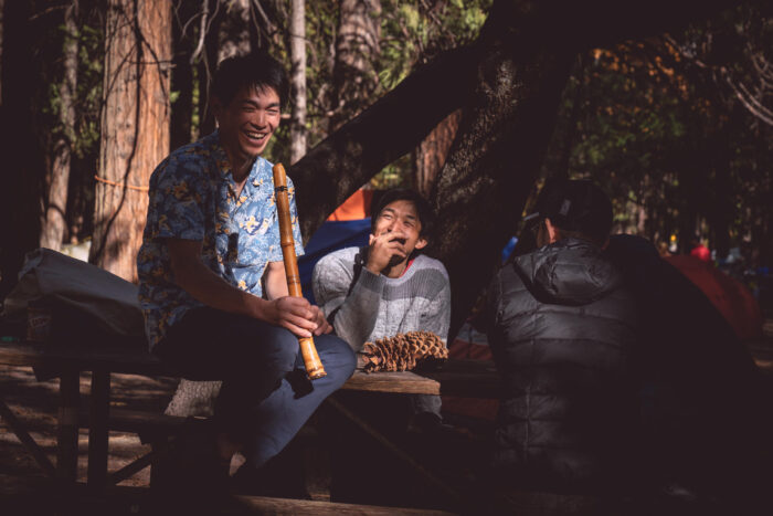 Kurakami plays the flute on a wooden table with some friends at Camp 4 under the pine trees