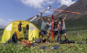Four skiers in shorts and t-shirts by a tent on a grassy meadow.