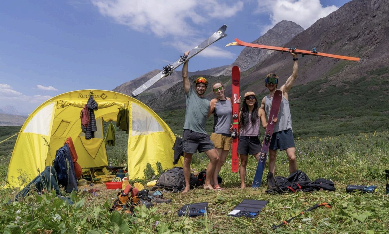 Four skiers in shorts and t-shirts by a tent on a grassy meadow.