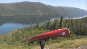 A man carrying a canoe up a hill, lake and mountains in the background.