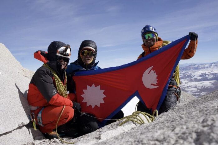 Three Sherpa climbers on a rocky summmit holding a Nepali flag.