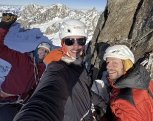 Three climbers on a belay station in a north face of the Alps, in the evening sun.