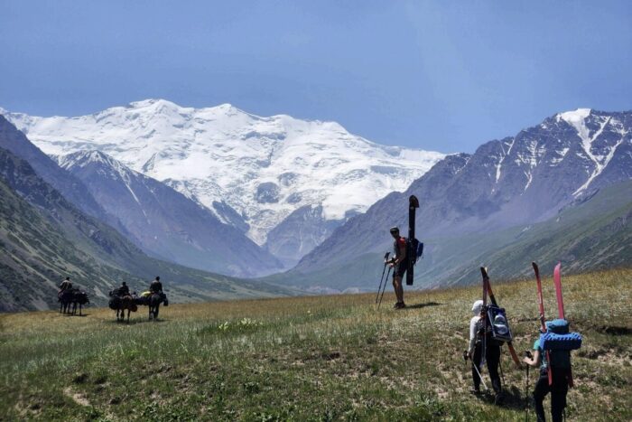 Climbers with skis on their backpacks walk on grassy meadows toward a snowy mountain range seen in background.