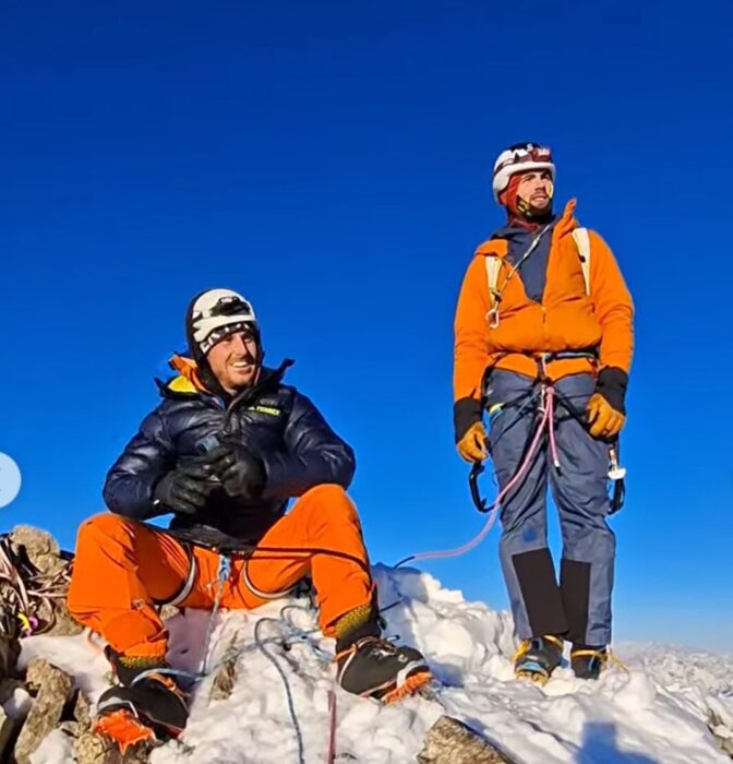 Two climbers on a mountain top, still roped up, in the sun. 