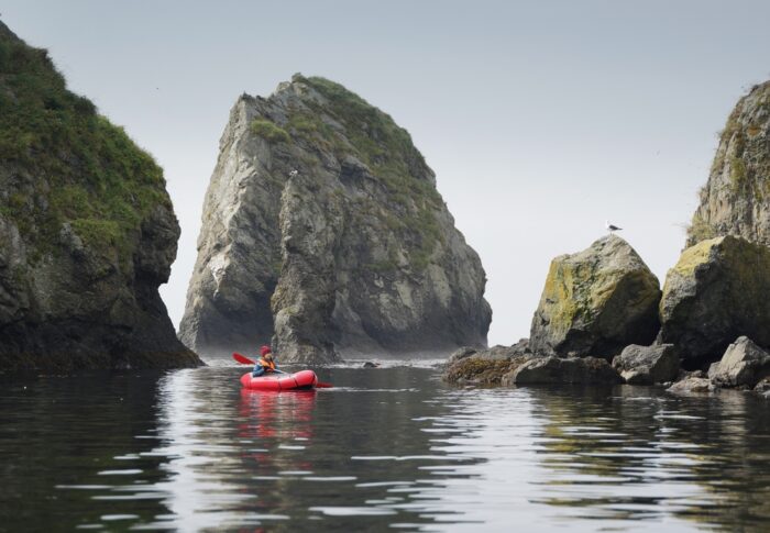 A solo pack rafter is pictured paddling on water surrounded by cliffs