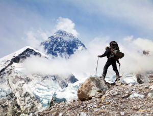 A man with a heavy backpack stands on a rock; in front of him, Everest's summit emerges from the clouds.