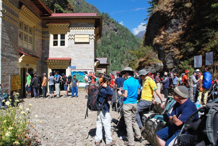 Trekkers stand causally soe steps away from a stone office house, hills covered in forests in background.
